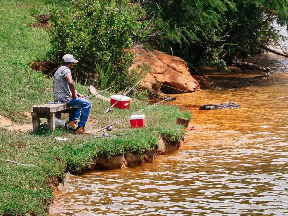 Pescador sentado no barranco do rio, com duas varas montadas, pescando de um jeito simples e barato